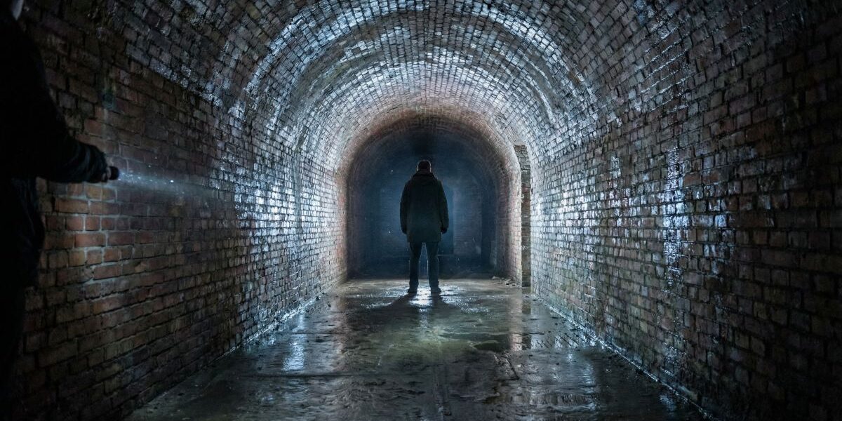 Ceglana podziemna tunelowa korytarz z kałużami i sylwetką człowieka oświetlaną latarką – tajemnicze miejsce w Wielkopolsce.