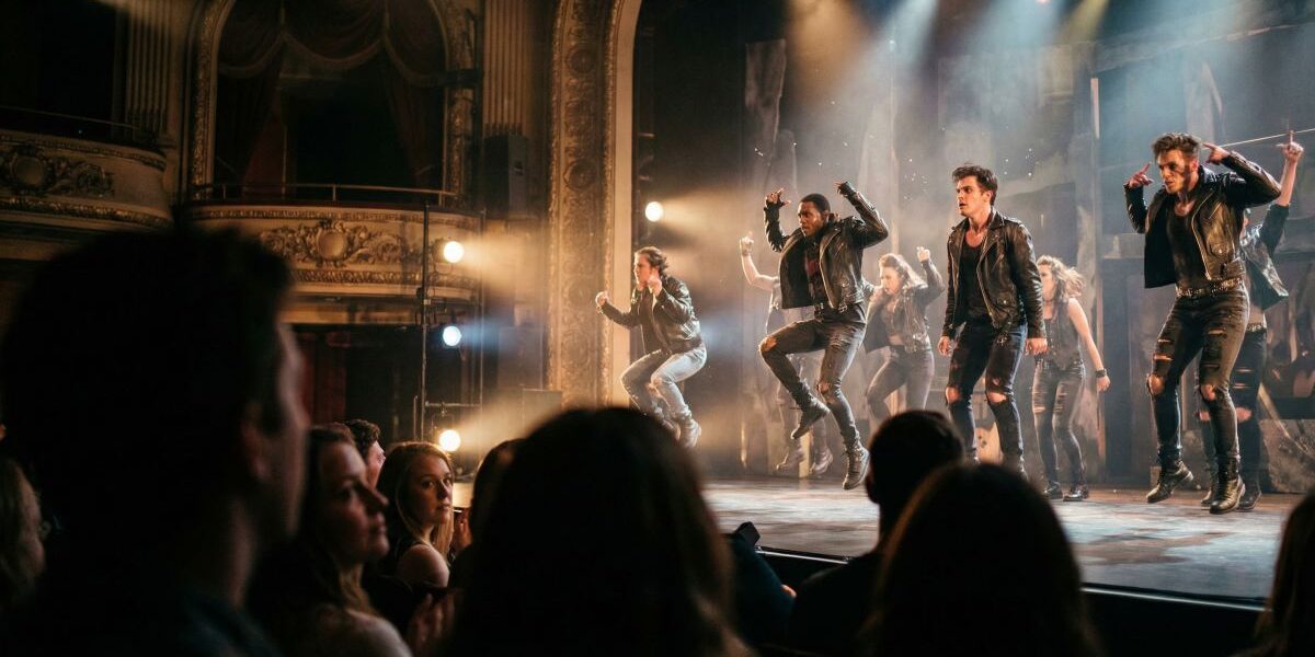 Dynamiczna scena teatralna: grupa tancerzy i aktorów w skórzanych strojach wykonuje układ na oświetlonej scenie, a widownia ogląda spektakl.